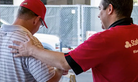 A photo shows a State Farm agent in a red shirt with a State Farm logo. He is patting a customer on the back. The customer is on the left and is wearing glasses, a red cap and a striped shirt. The photo is shot from behind.