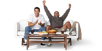 Two roommates cheer while sitting on their white couch. Snacks sit on the coffee table with two soda-filled glasses on either end.