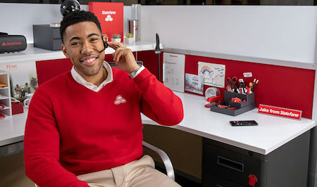 Image is of a young Black man, wearing a red sweater with the State Farm logo, a white shirt and khaki pants. He’s seated at a desk with a nameplate that says, “Jake from State Farm.” He’s wearing a headset with his left hand raised and fingertips touching the headset, indicating he’s in a call. He smiles at the camera.