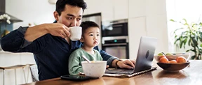 A young Asian American man sits before a laptop with a young male child sits on his lap at a kitchen table. The father, wearing a navy shirt, sips coffee from a white mug. His son wears a light green sweatshirt.