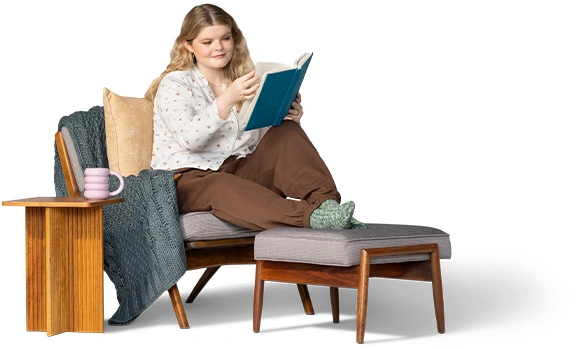 Una mujer está sentada leyendo en una silla cómoda, inclinada hacia adelante con los pies en calcetas sobre una otomana.