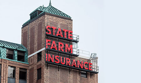 A photo of the State Farm Headquarters in Bloomington, IL. It’s a detail of the top of the brown-brick building. It has a teal green roof and features large, red neon letters that spell out “State Farm Insurance.” There is black railing in front of the neon site. 