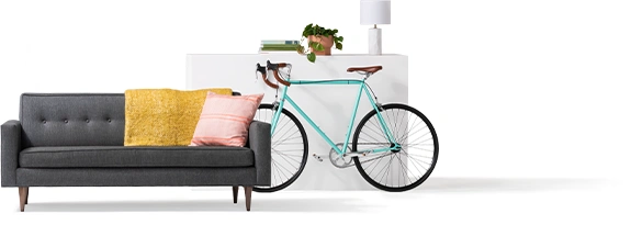 Hay dos almohadas sobre un sofá negro. Hay una bicicleta azul claro apoyada contra un baúl que tiene encima una lámpara y una planta.