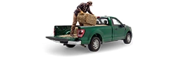 A bearded man in ballcap, flannel shirt and work pants prepares to throw a hay bale out of the back of a dark green pickup.