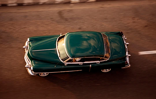 Vista aérea de lo que parece ser un Cadillac El Dorado verde esmeralda mientras avanza por la carretera.