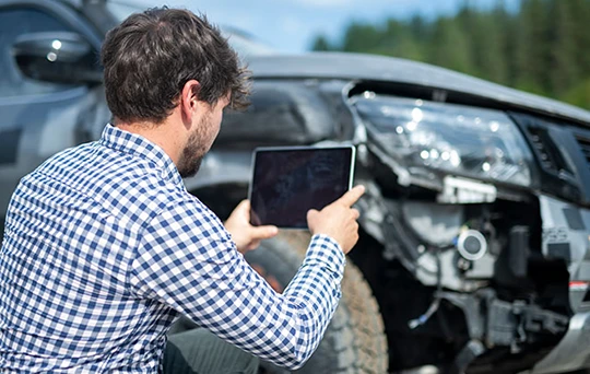 A man in a checkered shirt takes a picture of his vehicle’s front-end damage.