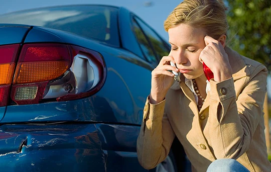 Listening to a phone call, a dejected woman in a tan overcoat kneels next to her damaged back bumper.
