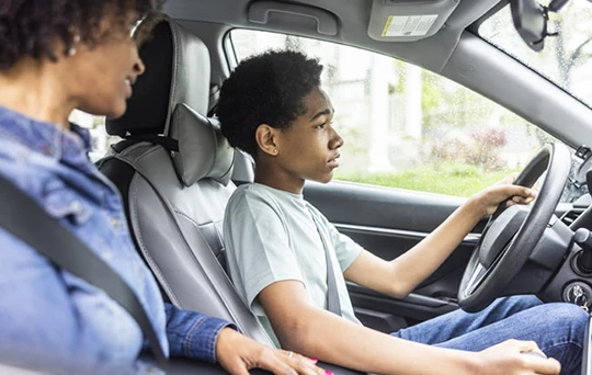 Una madre observa a su joven hijo, sentado en el asiento del conductor, mantener la vista fija en el camino.