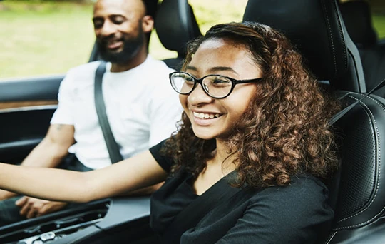 A young lady in glasses beams behind the wheel as her dad monitors her driving from the passenger seat.