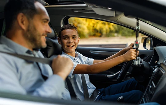 Before starting the car, a young driver smiles at his dad in the passenger seat.