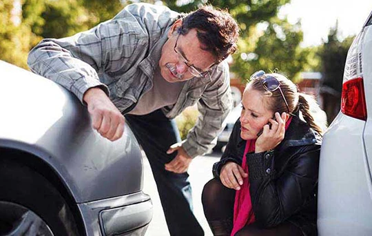 A man inspects damage to the front passenger side while a woman kneels nearby on her smartphone.