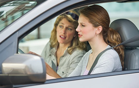 Una madre se inclina desde el lado del pasajero para revisar el espejo retrovisor mientras que su hija mantiene la vista en la carretera.