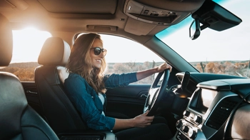Behind the steering wheel, a smiling woman in a blue blouse keeps her eyes on the road.