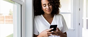A dark-haired woman, wearing a white top, looks at her mobile phone, while leaning against a wall next to a window.