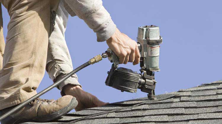 Man on roof fixing shingles with a nail gun.